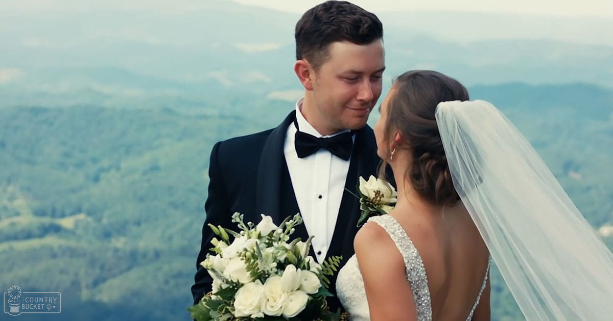 A young adult male singer in a black tuxedo and a young adult female wearing a white wedding dress with a long veil, holding a bouquet of white flowers against an overcast mountainous backdrop.
