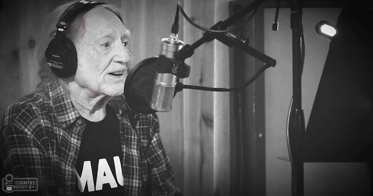 A middle-aged adult male wearing a plaid shirt and black t-shirt with 'MAL' written on it, seated in a professional recording studio setup.