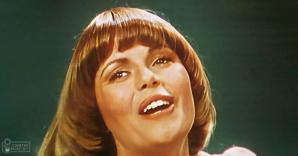 A young adult female indoors against a dark green backdrop, wearing a light-colored sleeveless top or dress under dim evening lighting.