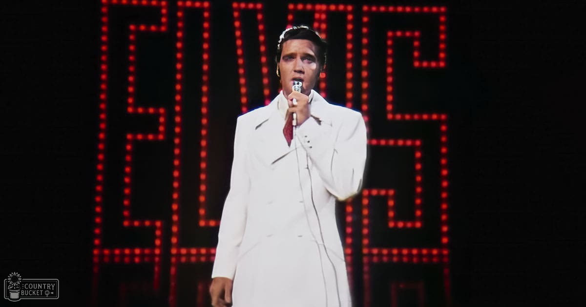 An adult male performer in a long white trench coat with a red tie, holding a microphone at an indoor Elvis tribute event.