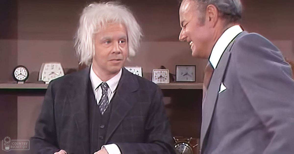 Middle-aged adult male wearing a black three-piece suit with a patterned tie, standing indoors near a vintage-style shelf holding clocks and framed pictures.