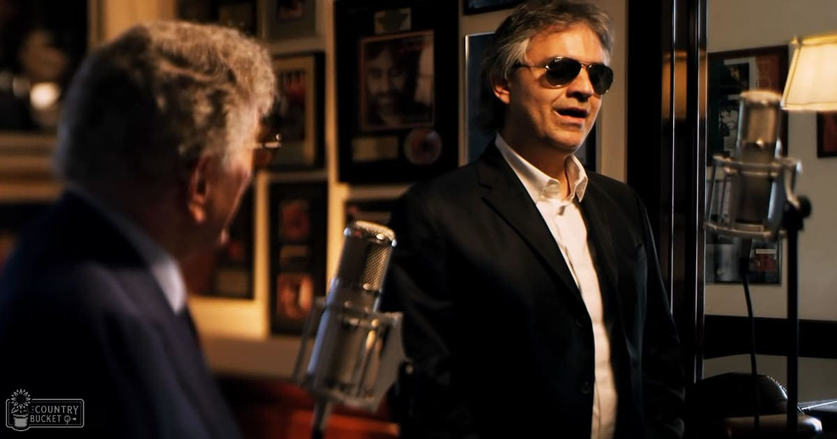 A middle-aged man in a dark suit jacket stands in a recording studio with another middle-aged individual wearing sunglasses and a blazer, surrounded by professional equipment.