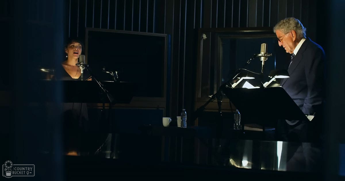 A musical performance in a recording studio with two adults, one wearing a dark top and another in a suit reading sheet music.