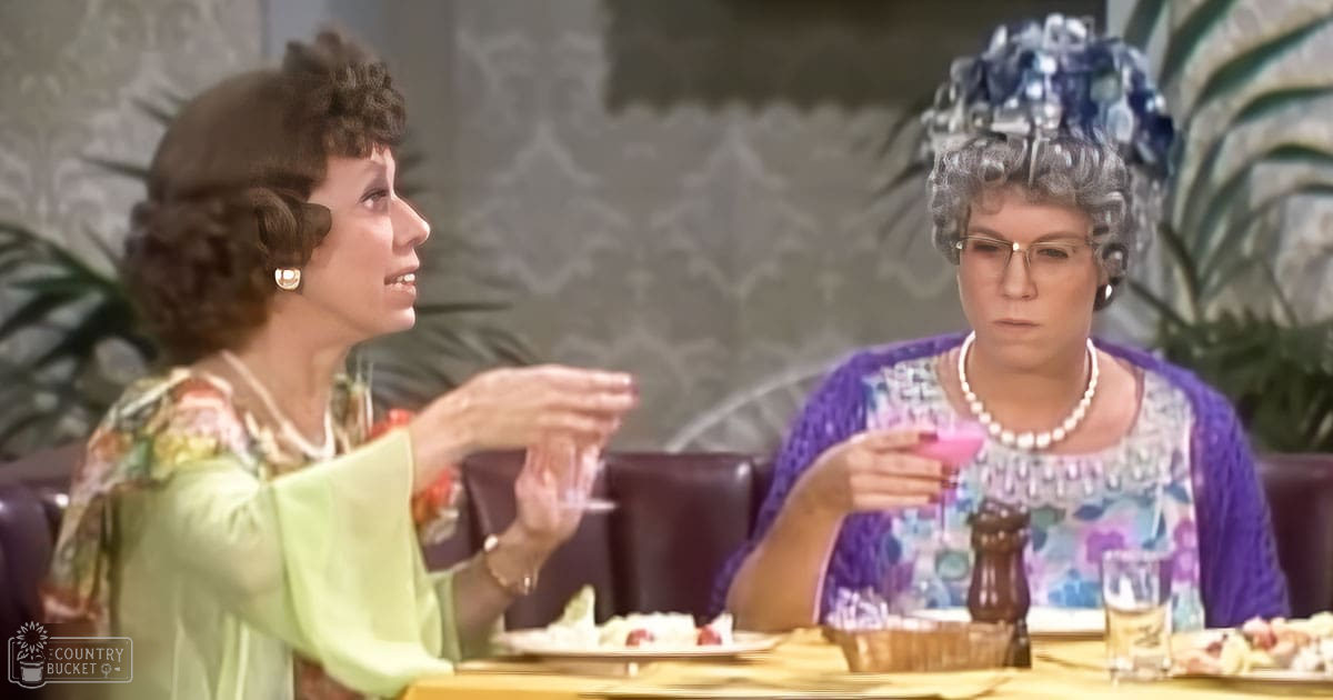 A middle-aged female wearing a light green, floral-patterned blouse with pearl earrings sits at a wooden table with yellow cloth, alongside an older adult female in a purple cardigan and colorful blouse. The scene is set indoors with ornate wallpaper and dark leather chairs.