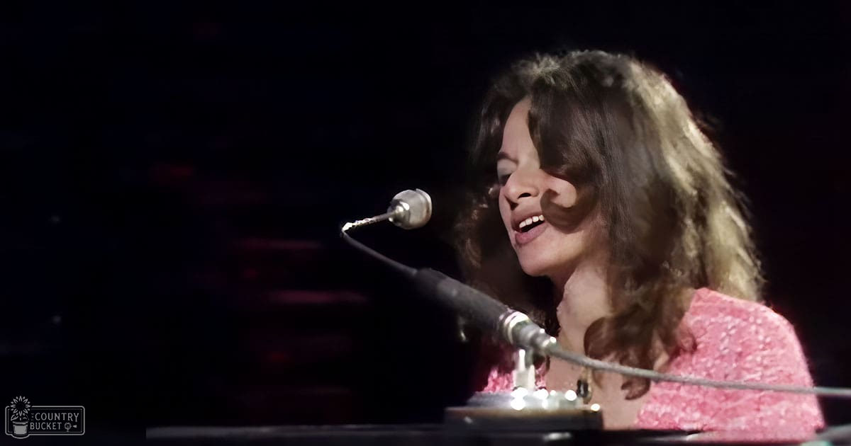 A young adult female in a loose-fitting, pink lace top performs on stage at a concert or live music event with a microphone stand and pedalboard visible.