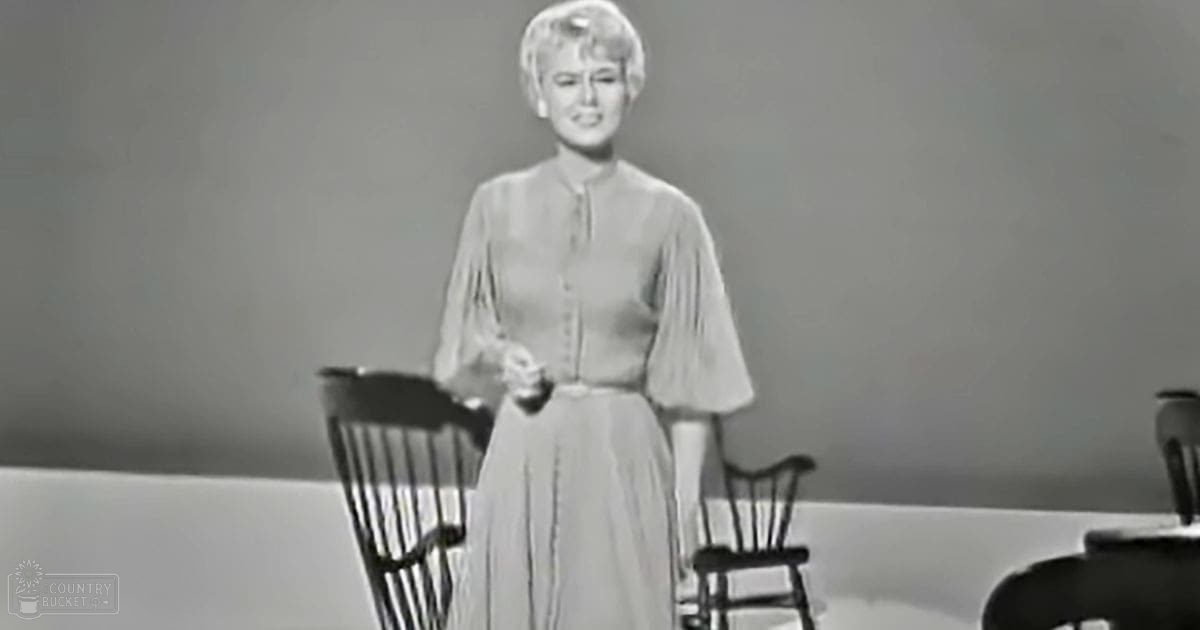 A young adult female in a light gray dress with puffed sleeves stands indoors against a plain background.