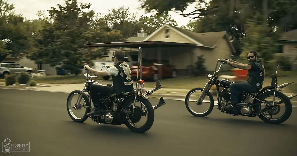 Two adult males riding motorcycles in a suburban neighborhood during daytime with earthy tones dominating the scene.