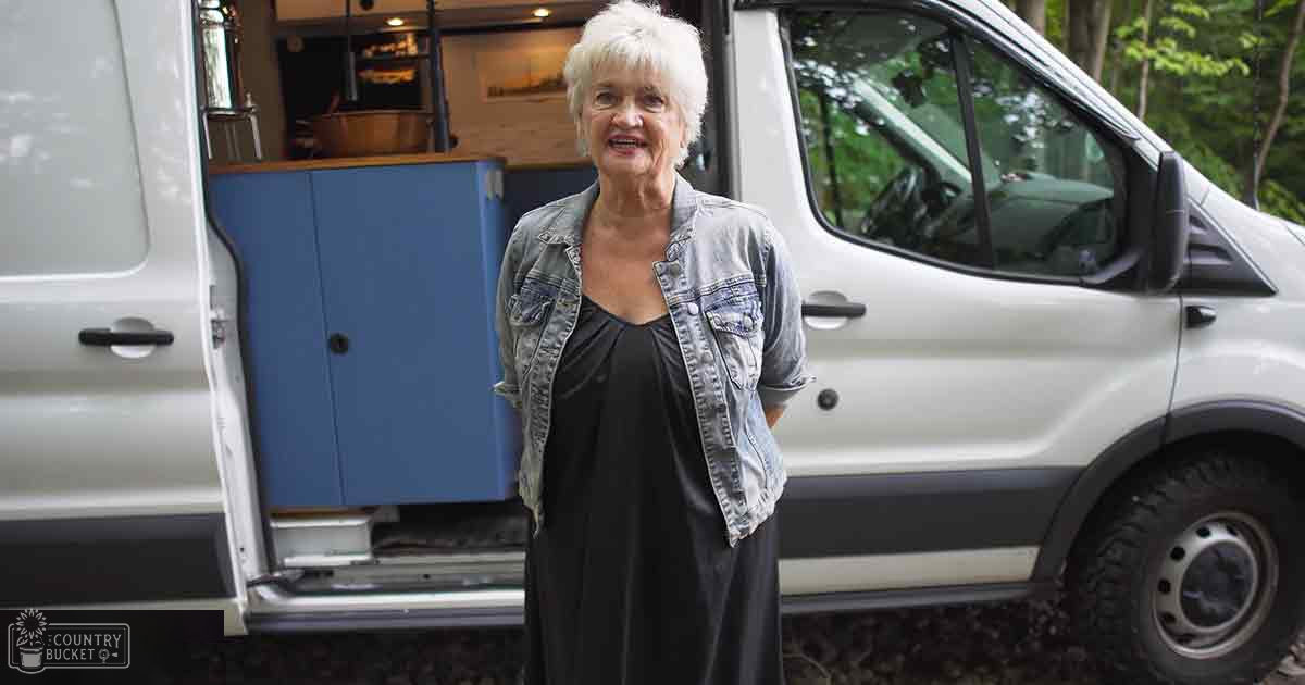An older adult female wearing a denim jacket and black dress, standing outdoors near a white van with open doors revealing its interior in a rural or forested area during spring or summer day.