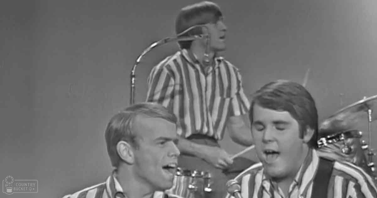 Musical performance scene from the 1960s with young adult males in vertically striped shirts against a simple stage backdrop with vintage microphone and guitar straps.