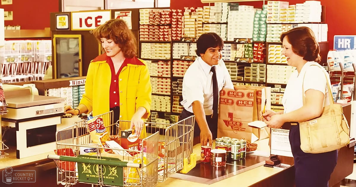 A cashier at H-E-B wearing a yellow jacket over a red shirt, interacting with customers in the store. A shopping cart filled with groceries and shelves stocked with products are visible behind her.