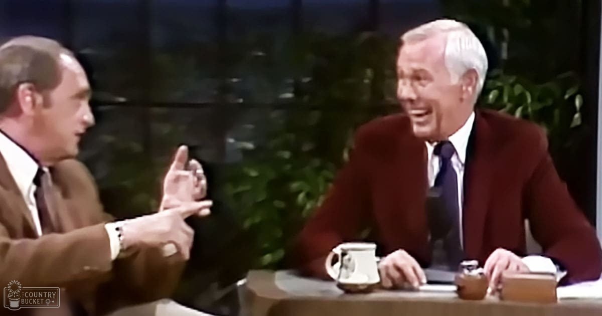 An adult male talk show host and an older adult male seated at a table with a mug, indoor studio setting with plants and artificial lighting.