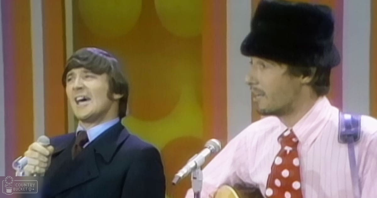 A mid-20th century musical performance scene with two young adult men in formal attire, one holding a microphone and the other wearing a fur hat with a guitar strap, set against an indoor studio backdrop with circular yellow-orange shapes.