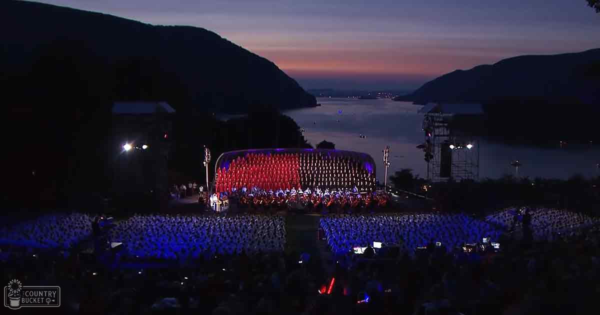An outdoor performance at dusk with a scenic backdrop of mountains and water with audience members in white shirts and performers in red and blue costumes.