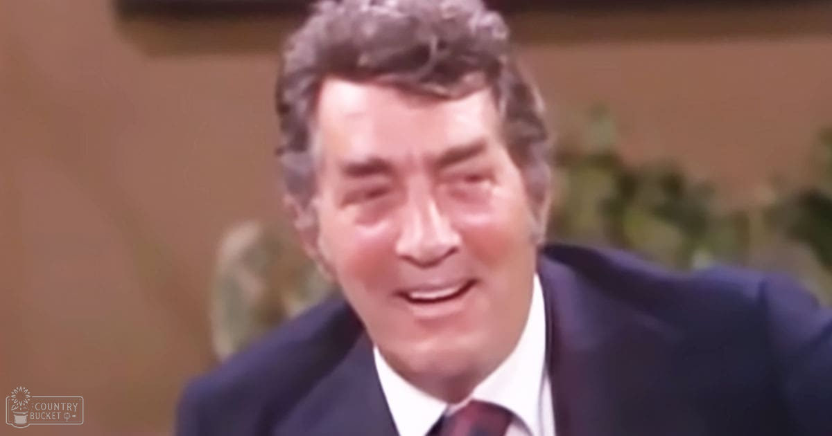 A middle-aged adult male dressed formally in a dark blue suit with a white shirt and red tie, set against an indoor backdrop with blurred greenery and warm lighting.