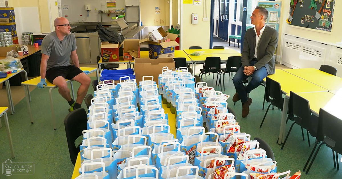 Two adult males in a classroom setting, one wearing a gray T-shirt and black shorts, the other in a dark gray blazer over a white shirt with brown boots. Yellow tables and various food items packed into blue bags are visible.