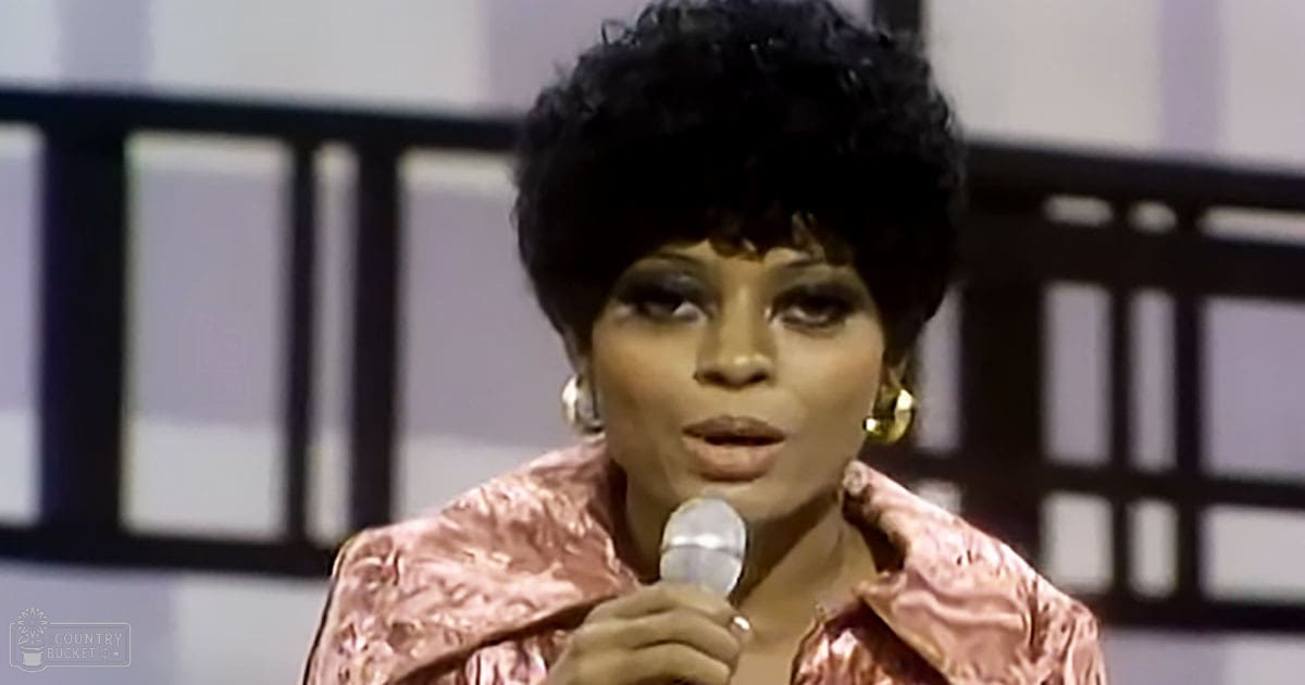 A woman performs on stage, wearing a shiny pink satin-like jacket with puffed sleeves and high collar, in front of geometric-patterned backdrop under even studio lighting.