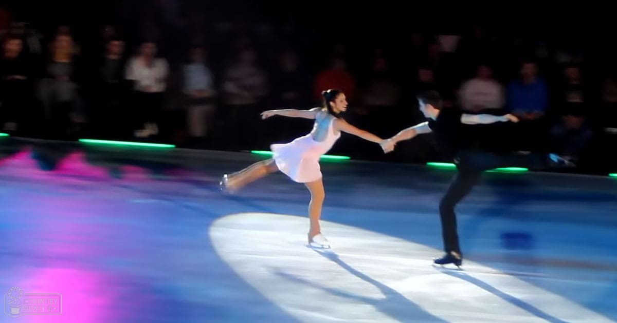 A figure skating pair performing on an indoor ice rink with spectators seated in the background.