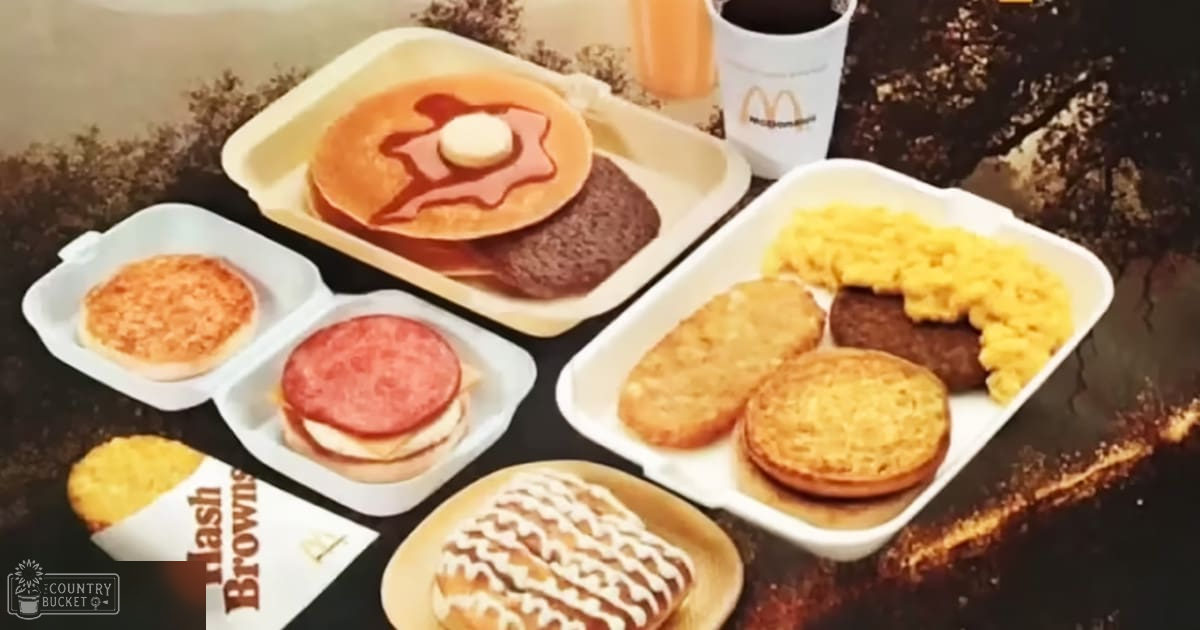 McDonald's breakfast meal with hash browns, sausage patties, scrambled eggs, pancakes topped with syrup and butter, bacon, donuts with icing, and a cup of coffee or tea on an indoor table.