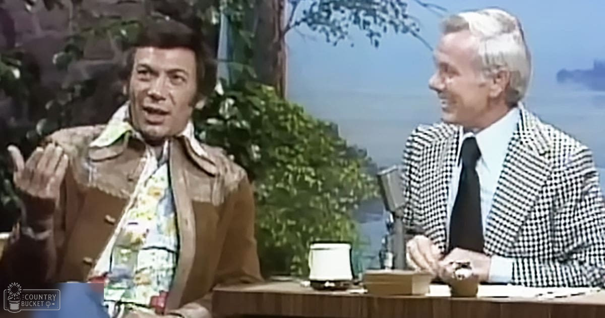 Talk show host and guest on an outdoor studio set with lush greenery and a body of water in the distance, suggesting spring or summer.