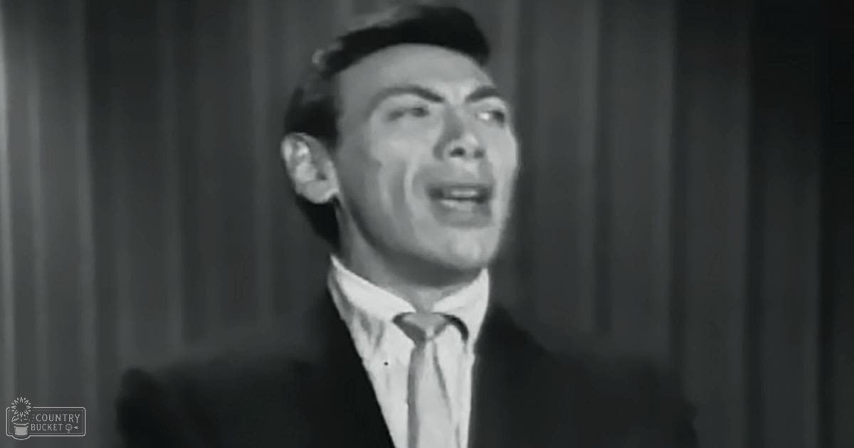 A young adult male in a tailored dark suit jacket with a tie, white collared shirt, and a hat on his head stands indoors under stage lighting.