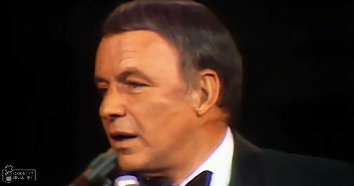 A middle-aged man wearing a dark suit jacket, white shirt, and black bow tie stands indoors with a microphone near his mouth, suggesting a performance or recording session.