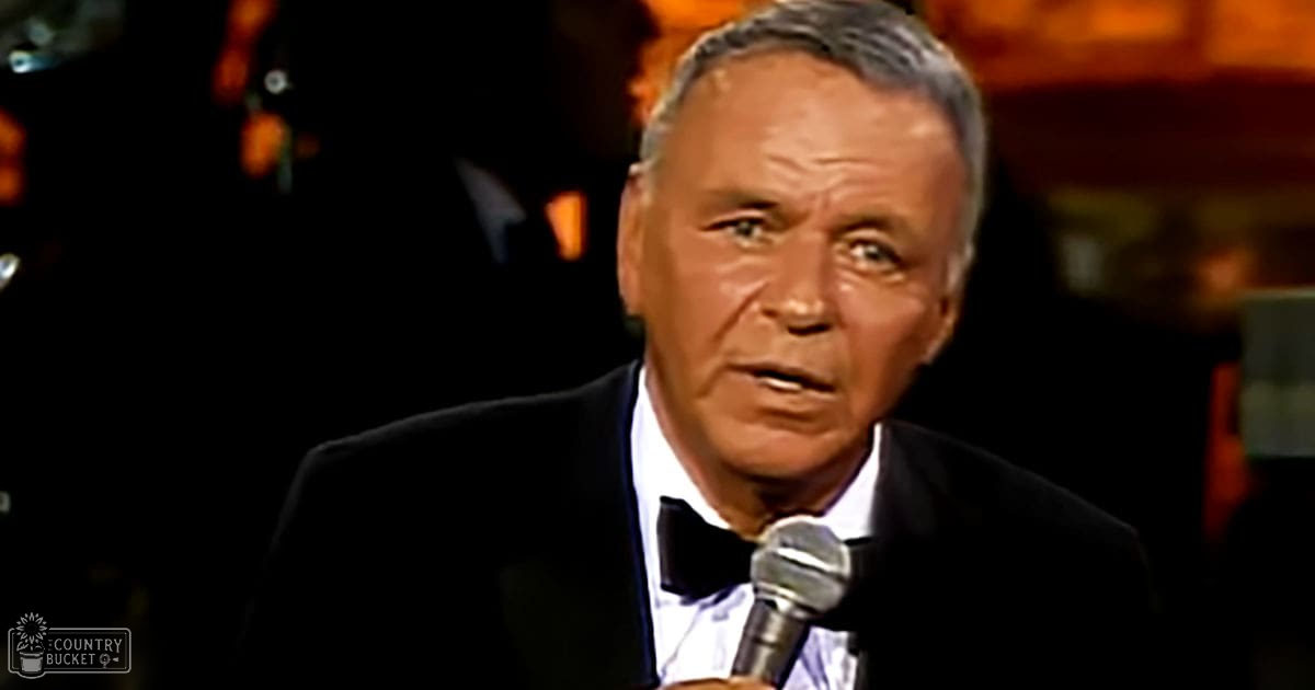A middle-aged man in a black tuxedo with a white shirt and black bow tie, holding a microphone on stage under artificial lighting at night.