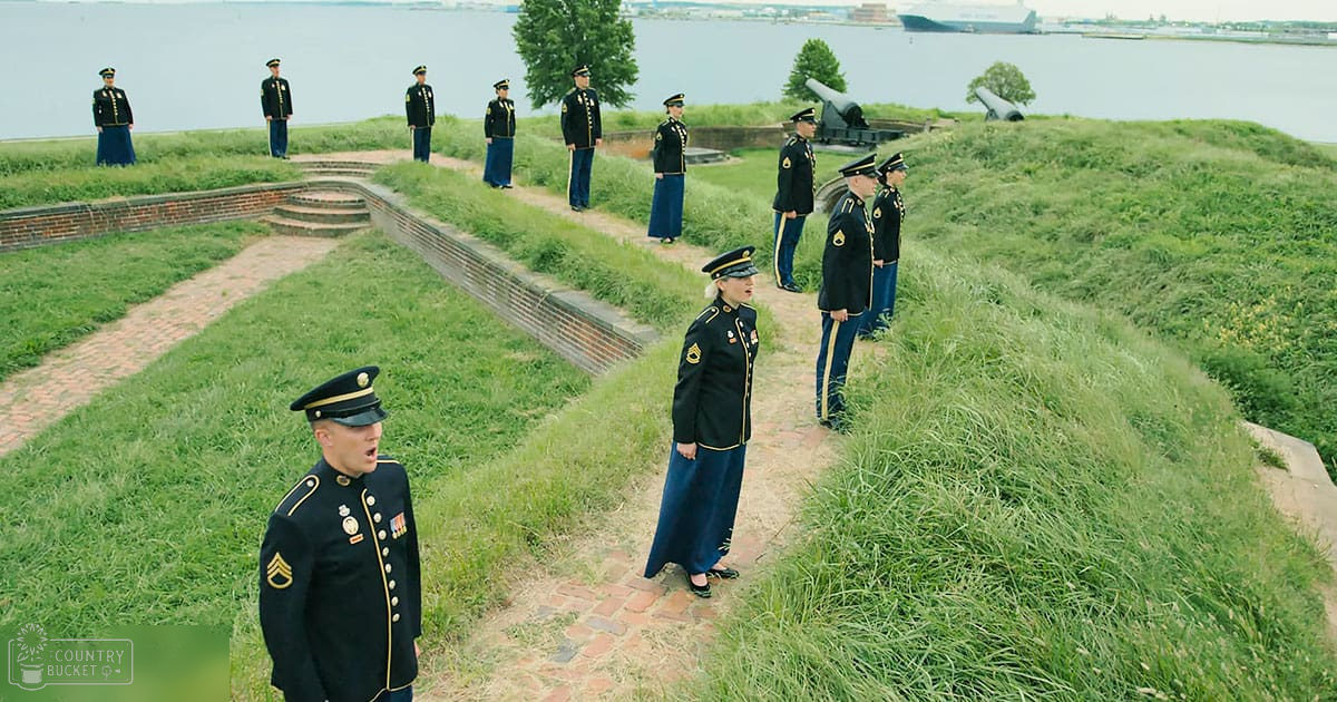 A group of uniformed individuals in a military setting at Fort McHenry, Baltimore, Maryland, with cannons and lush greenery surrounding the scene during daylight hours.