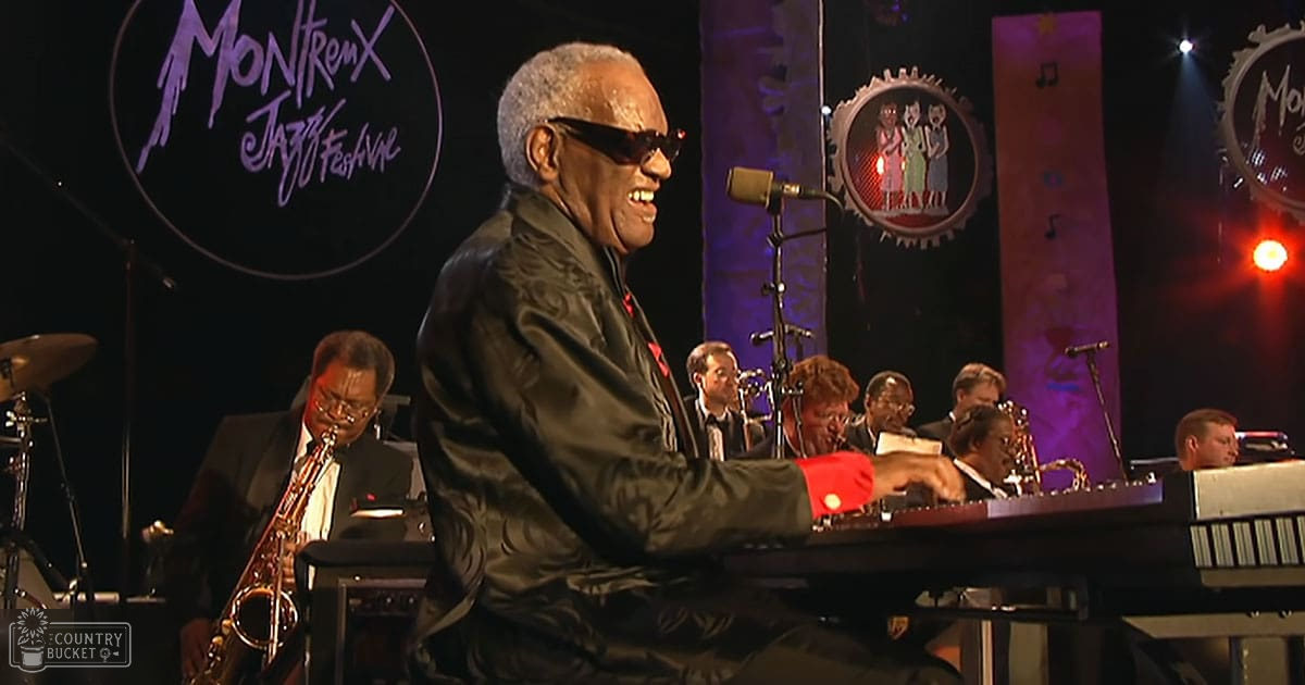 An older adult male performer at the Montreux Jazz Festival, wearing a black satin jacket with red accents and dark sunglasses; accompanied by other band members in formal attire under stage lighting.