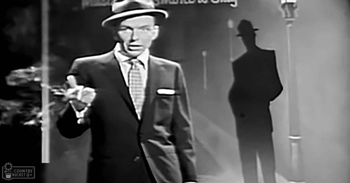 An adult male singer performing on stage with a formal suit, tie, and hat in a dimly lit indoor setting.