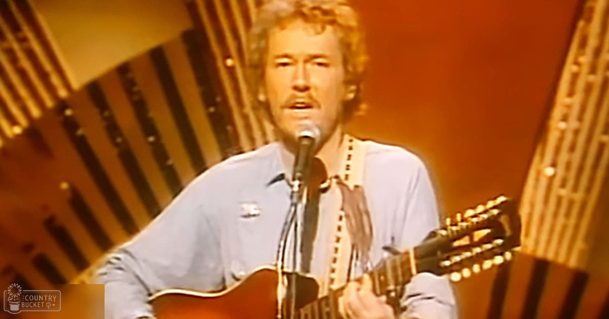 A middle-aged male singer performs on stage, wearing a light blue button-up shirt with white suspenders and holding an acoustic guitar.