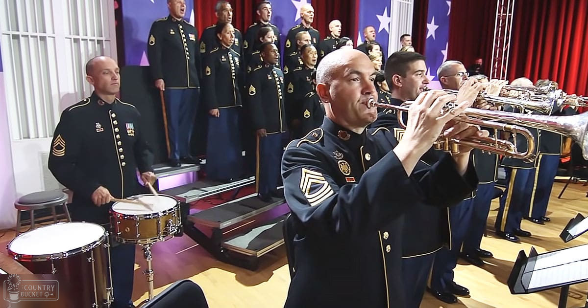 A trumpet player in a dark blue military-style uniform with gold trim and insignia stands at an indoor stage, surrounded by other adult male musicians in similar uniforms, against a backdrop of red curtains and American flags.