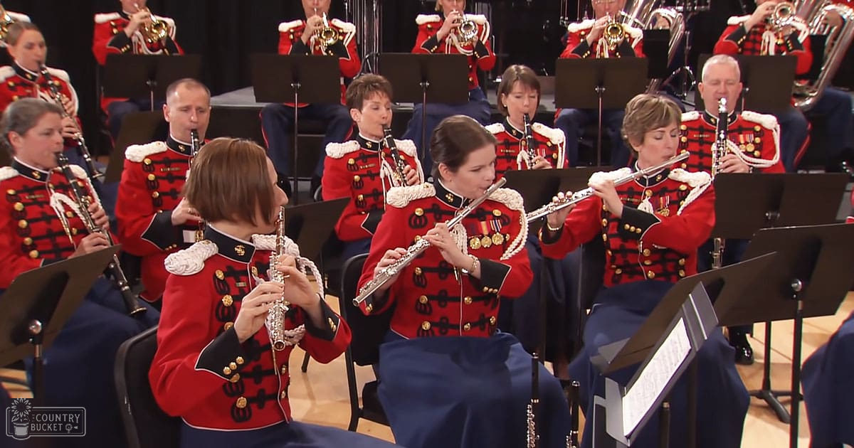 An adult female singer in a red jacket with gold buttons and white fur trim, black pants, holding an instrument at an indoor concert setting filled with musicians and brass instruments.