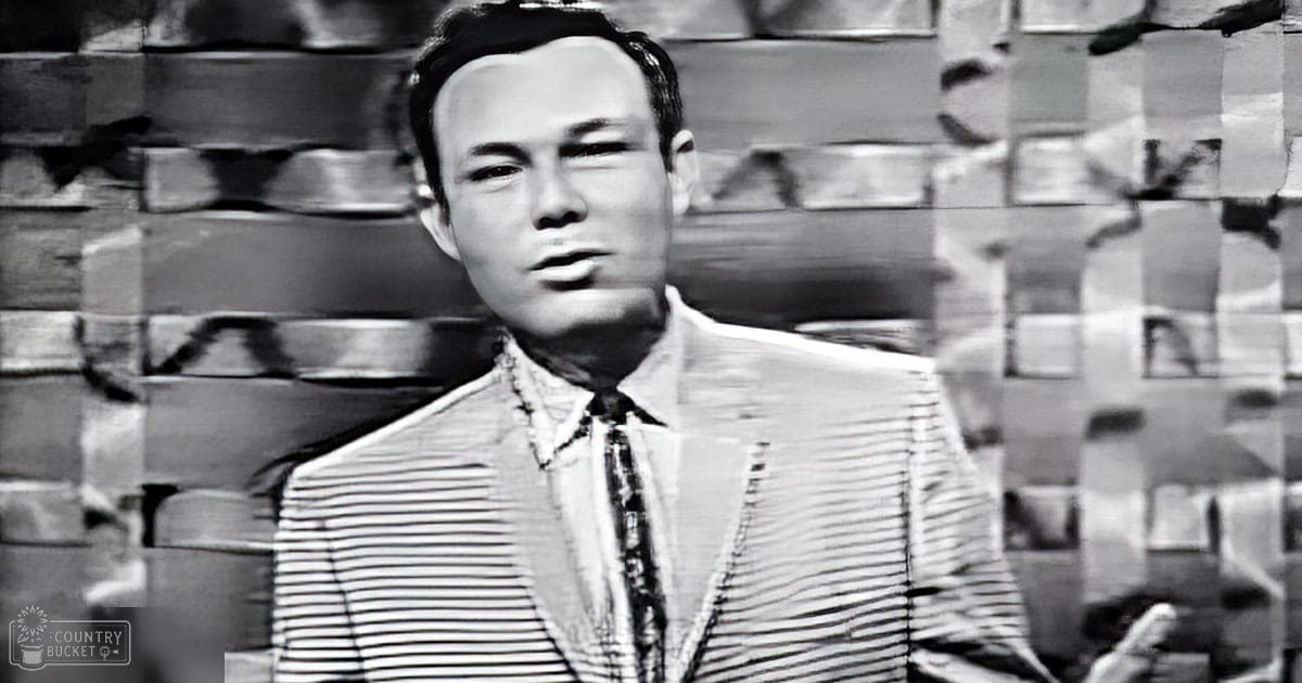 A young adult male in a striped suit jacket stands indoors against a textured brick wall with even artificial lighting.