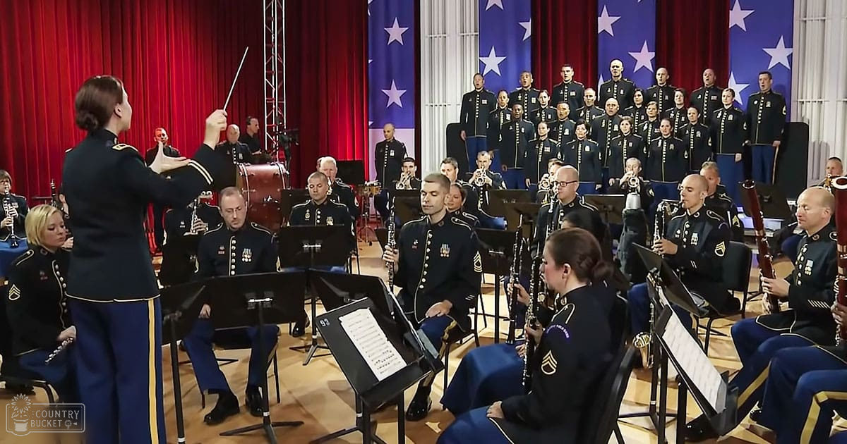 Adult female conductor leading an orchestra in a concert hall, surrounded by musicians and choir members wearing matching dark blue military-style uniforms with gold trim. Musical instruments like clarinets and flutes are visible, along with sheet music stands, red curtains as backdrop, American flags behind the choir.