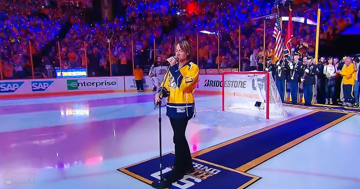A middle-aged male singer in a yellow and blue sports jacket with the number 7, performing at an ice hockey arena filled with a patriotic crowd.