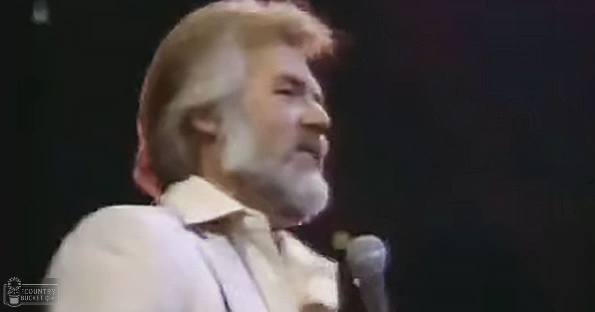 A middle-aged adult male performer wearing a white, collared shirt labeled 'Madly Odd' sings into a handheld microphone on stage in an indoor venue with dim lighting.