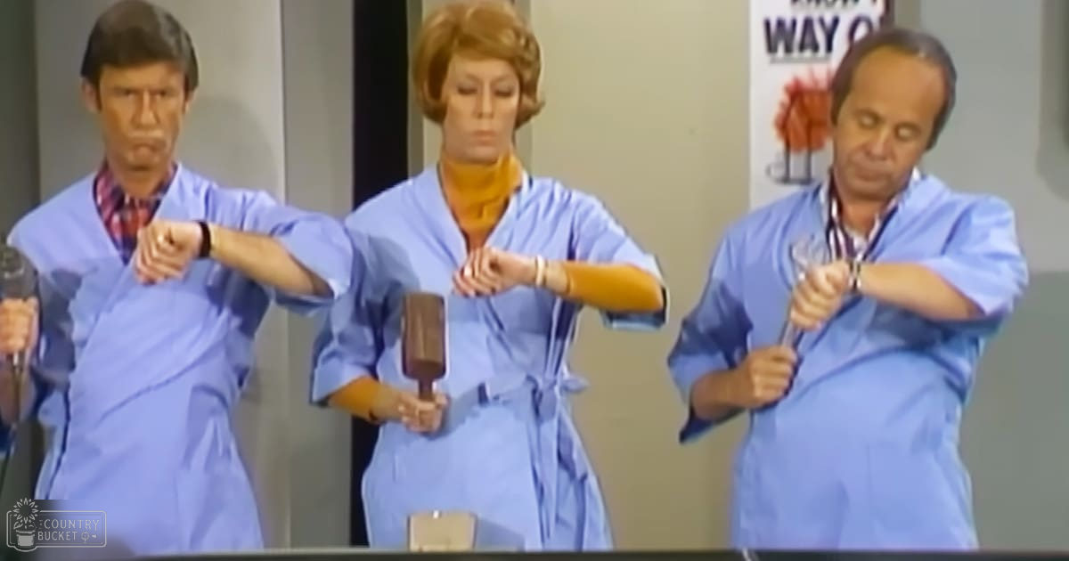 Medical or hospital environment scene from a television show or sketch comedy with adults wearing surgical gowns and holding props, set indoors with neutral-colored walls and a 'WAY OUT' sign.