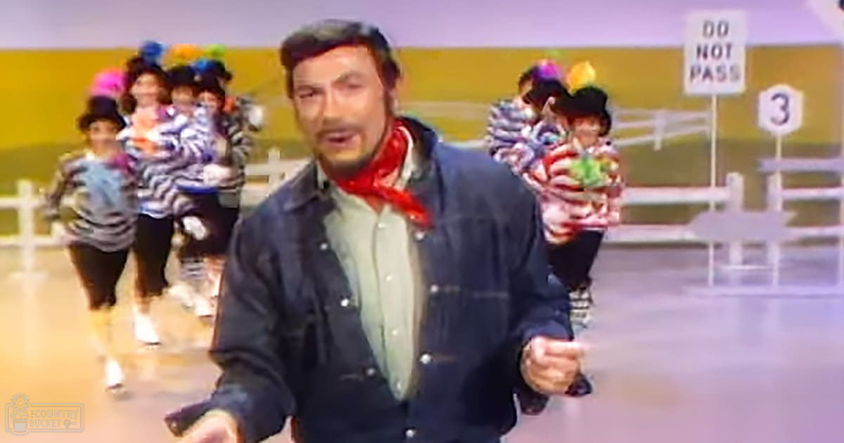 Multiple children in costumes, an adult male wearing a dark jacket with red scarf on an indoor stage or studio backdrop with road signs and fence.