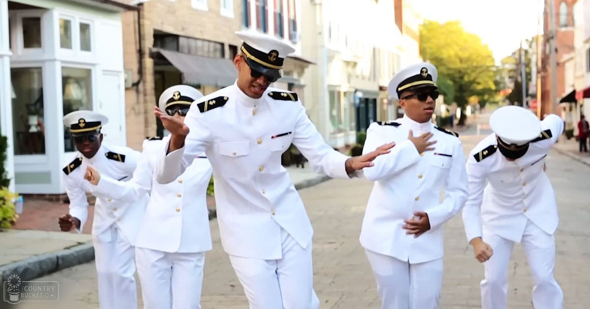 Young adults in uniform dancing on a sunny urban street with greenery along the sidewalk, suggesting spring or summer.