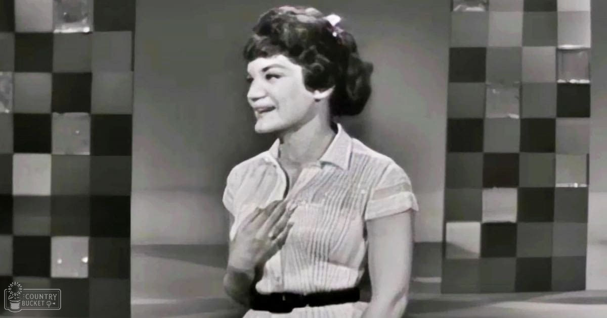 A young adult female wearing a short-sleeved, collared shirt with vertical stripes and a belt at the waist, standing against a checkered wall in an indoor setting.