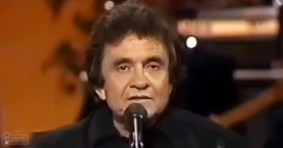 A middle-aged adult male wearing a dark-colored collared shirt or jacket with long sleeves is performing indoors, surrounded by musical instruments.