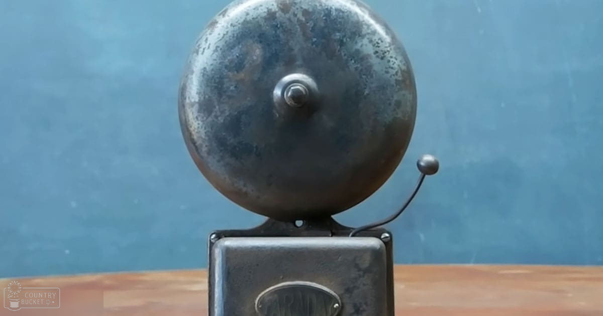 An old-fashioned metal bell with a worn appearance, mounted on a rectangular base with text reading 'ARM' against a plain blue indoor background.