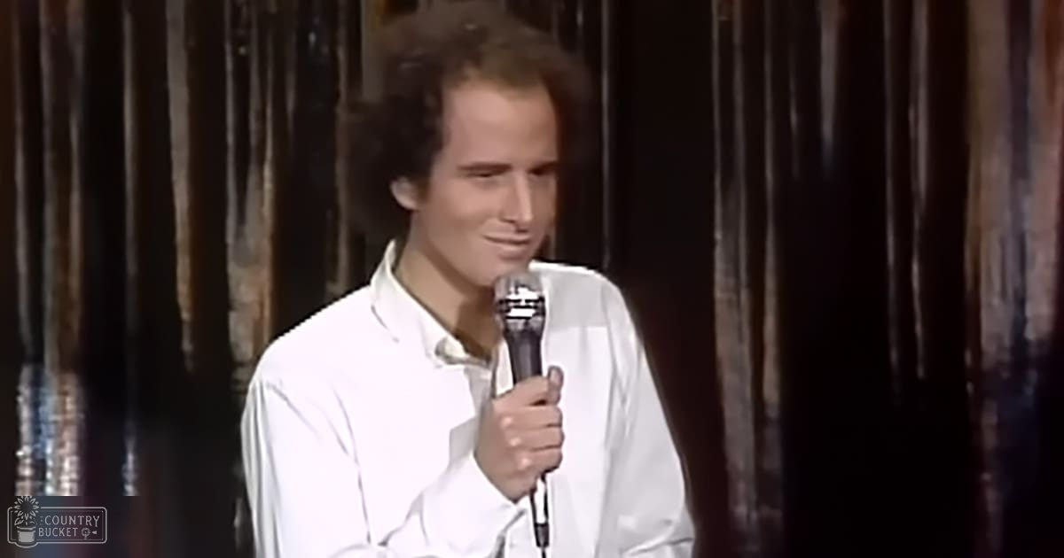 A young adult male performs stand-up comedy in a dimly lit indoor setting, wearing a white collared shirt with dark curtains behind him.