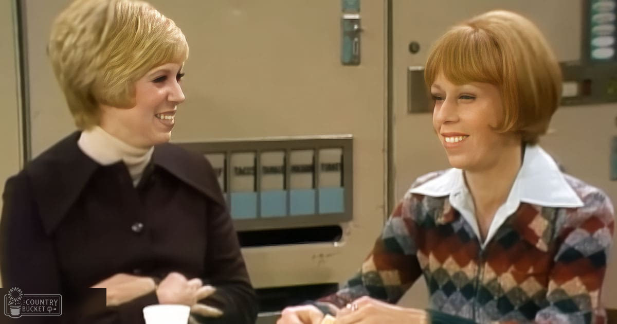 Two young adult women in a workplace setting, one wearing a dark-colored, oversized collared jacket and the other in a plaid zip-up sweater with long sleeves, holding coffee mugs. A vending machine is visible in the background.