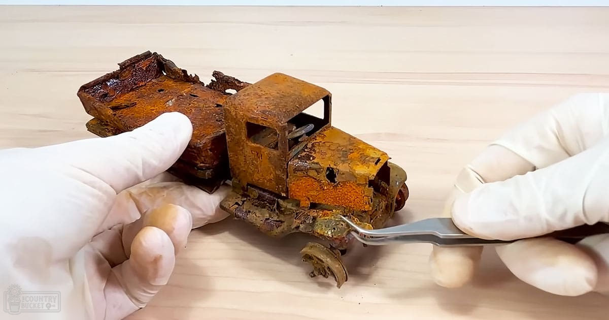 A close-up view of a rusted toy truck being held by hands wearing white gloves on an indoor light-colored wooden surface.
