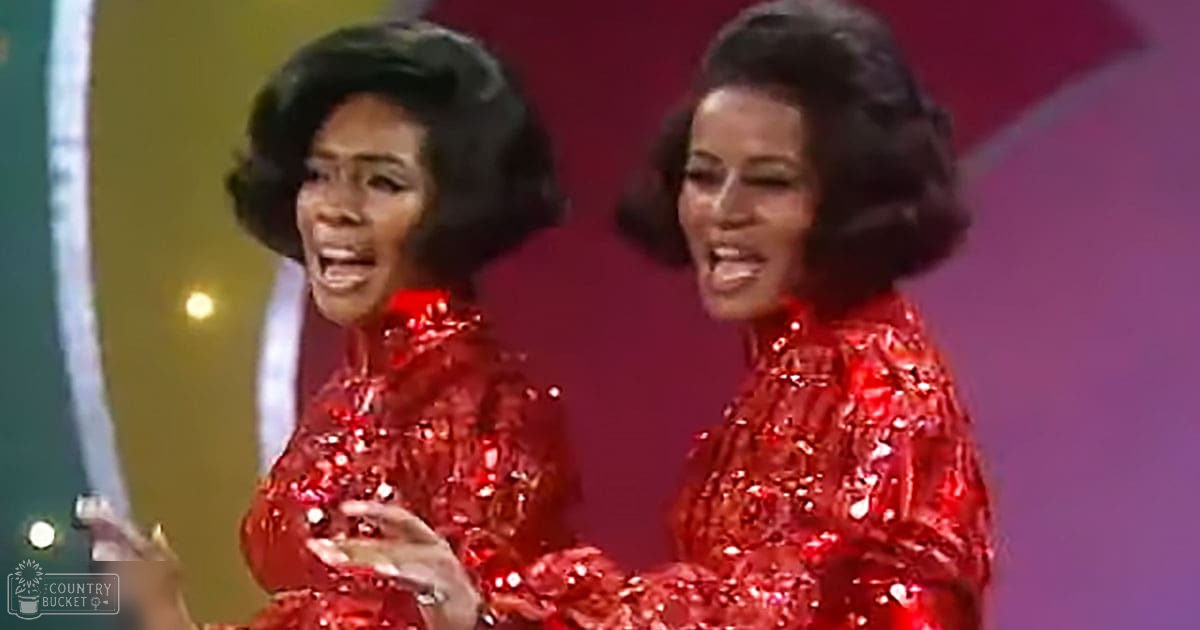 Two adult female performers on stage in red sequined dresses from the 1970s or 1980s, holding microphones against a vibrant backdrop of green, yellow, and purple colors.