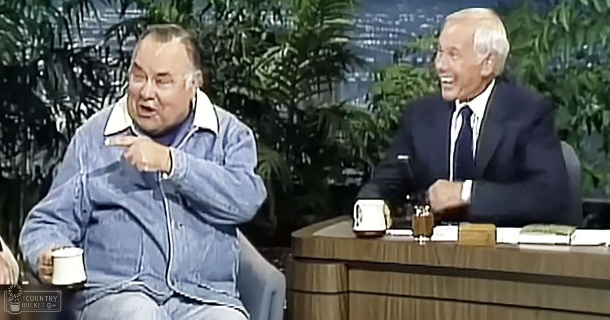 Two adults seated at a wooden desk with green plants and an urban night backdrop, suggesting a talk show environment.
