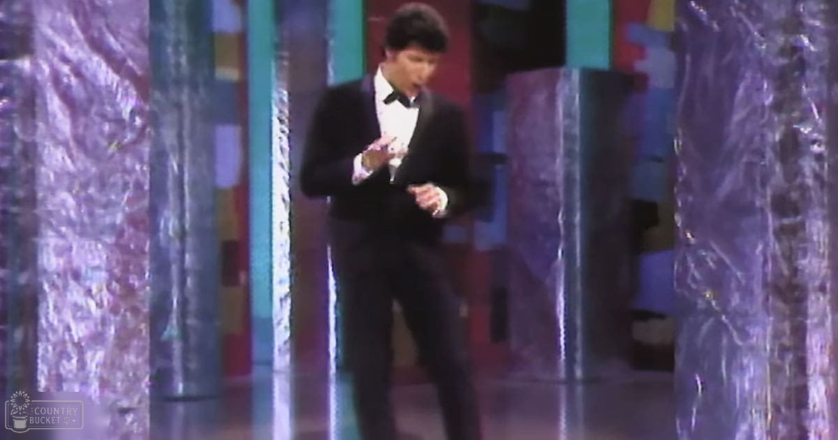 Young adult male wearing a formal black tuxedo with white shirt, holding a trophy on stage at an awards show.