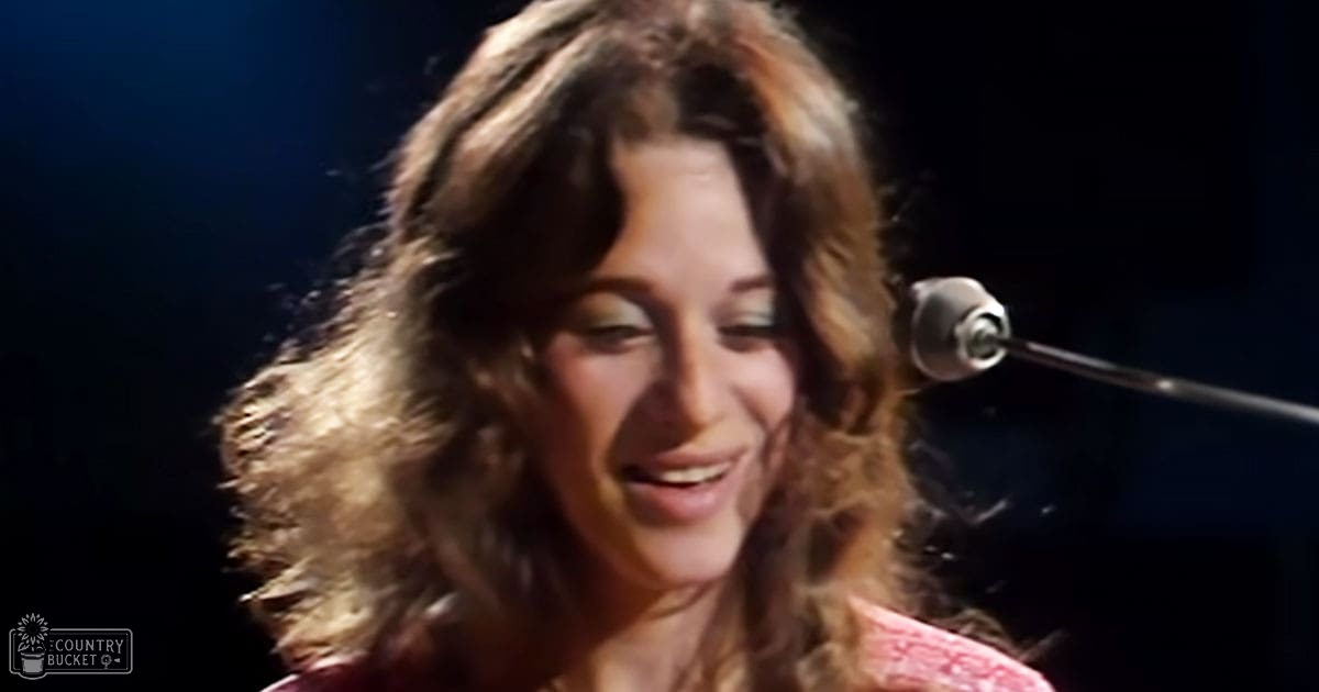 A young adult female stands indoors with a microphone stand, suggesting a performance or speech setting.