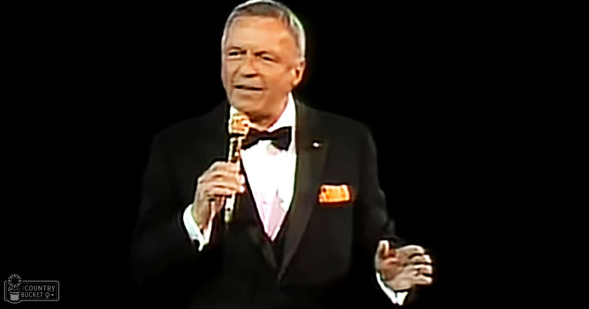 A middle-aged adult male singer wearing a classic mid-20th-century style tuxedo with a white dress shirt, black bow tie, and orange pocket square on stage.